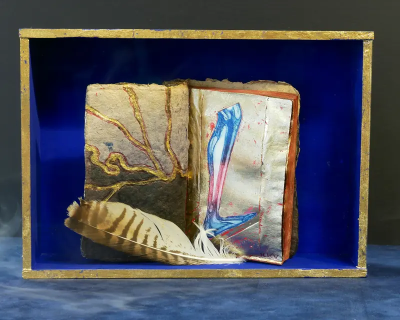 Objektkunst: ein bemaltes Buch mit Feder in einem blauen Kasten mit vergoldetem Rand